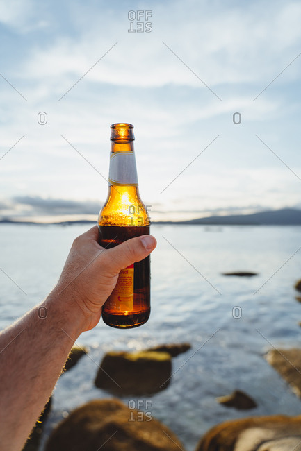 Crop traveling person holding shiny bottle of beer against sunlight resting on remote seashore