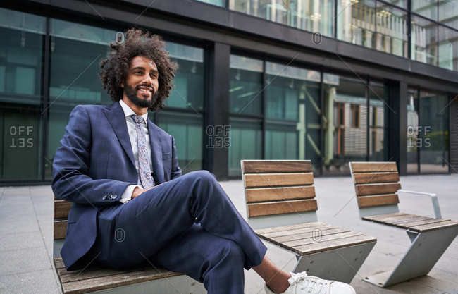 Optimistic and merry african american entrepreneur with beard sitting on bench with crossed legs and looking away