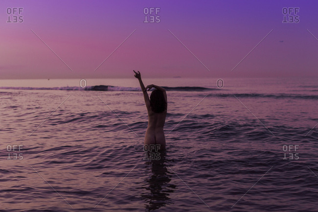 Back view of sexy woman with raised hands standing in water and enjoying landscape on gradient pink purple sky background