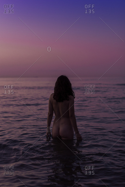 Back view of sexy woman with raised hands standing in water and enjoying landscape on gradient pink purple sky background