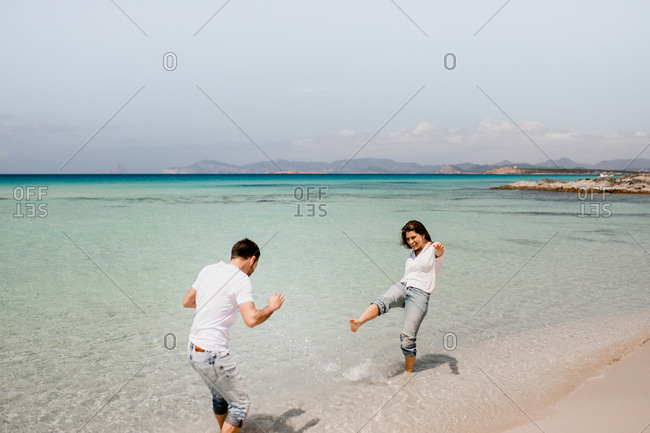 Playful man and woman in white shirts playing in shallow water of seashore in summer