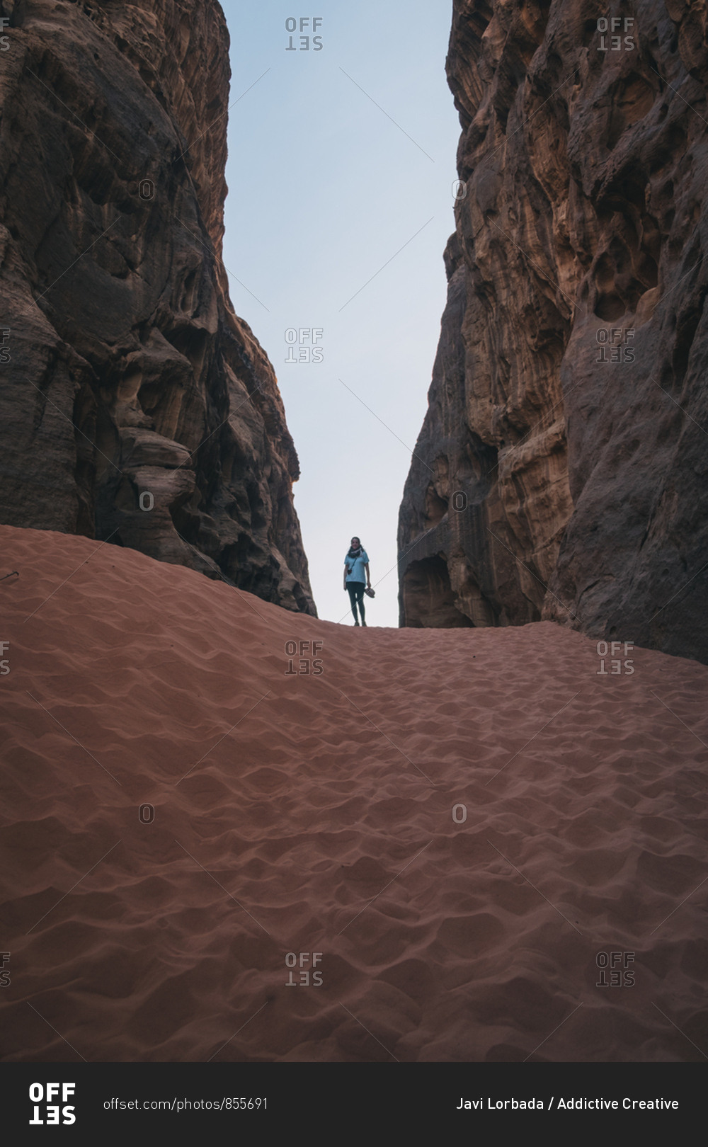 Anonymous female tourist standing on sand between rough stone walls against cloudless blue sky in ravine of wadi rum desert in jordan