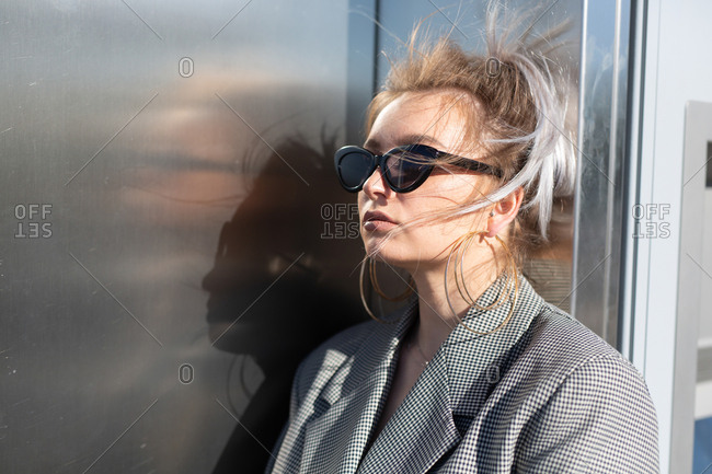 Fashion attractive woman with trendy hairstyle and sunglasses nearby dark shiny metal wall looking away