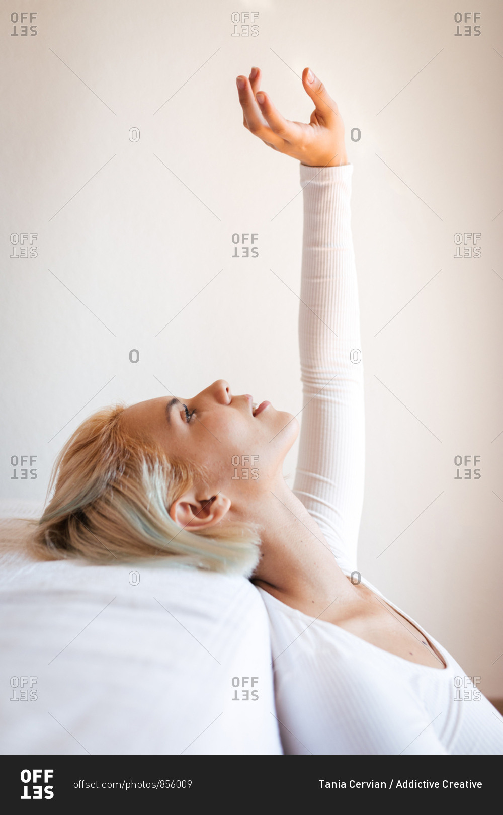 Side view of beautiful blond lady looking up and leaning on comfortable mattress against white wall at home