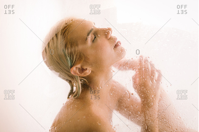 Side view of young female taking shower behind wet transparent partition in bathroom
