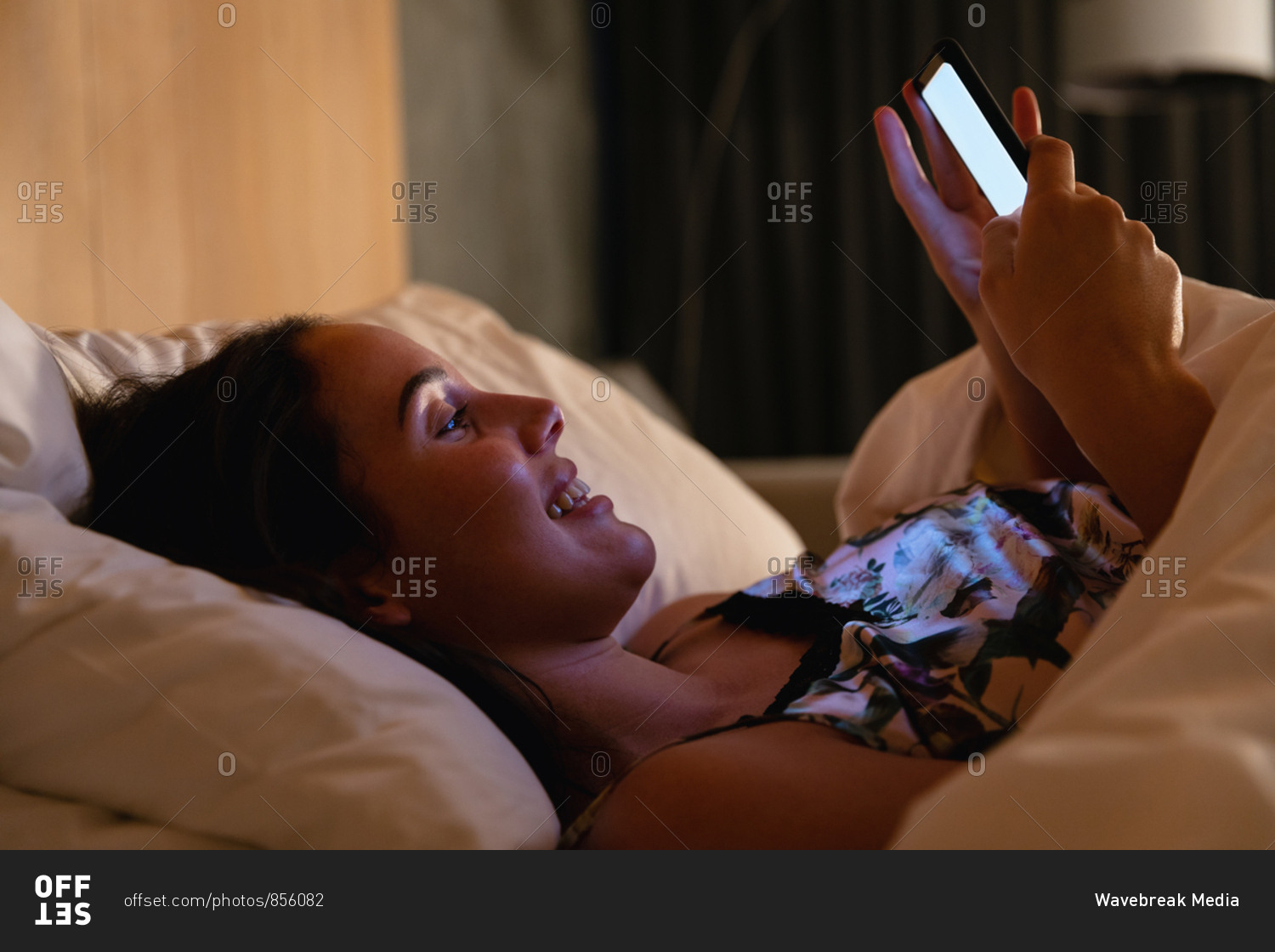 Side view close up of a smiling young Caucasian brunette woman lying on her back in bed using a smartphone