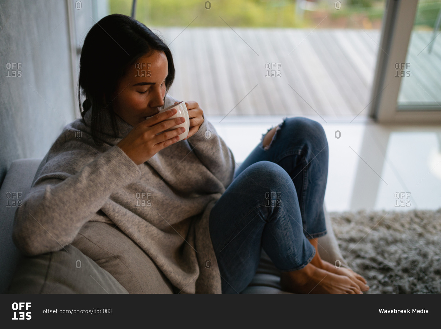 Elevated view of a young Caucasian brunette woman sitting on a sofa with her legs drawn up enjoying a cup of coffee