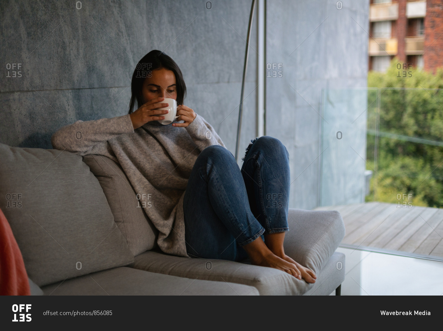 Front view of a young Caucasian brunette woman sitting on a sofa with her legs drawn up, enjoying a cup of coffee