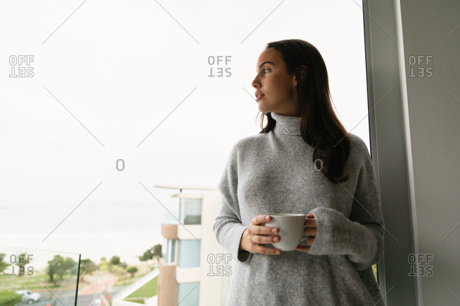Front view of a young Caucasian brunette woman wearing a grey turtleneck sweater, standing on a balcony holding a cup of coffee with her head turned to the side, looking away