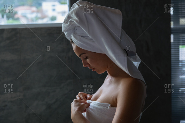 Side view close up of a young Caucasian woman wearing a bath towel and with her hair wrapped in a towel, standing in a modern bathroom looking down