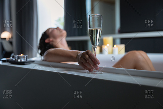 Side view of a young Caucasian brunette woman lying in a bath with a lit candles on the side, leaning back and holding a glass of champagne