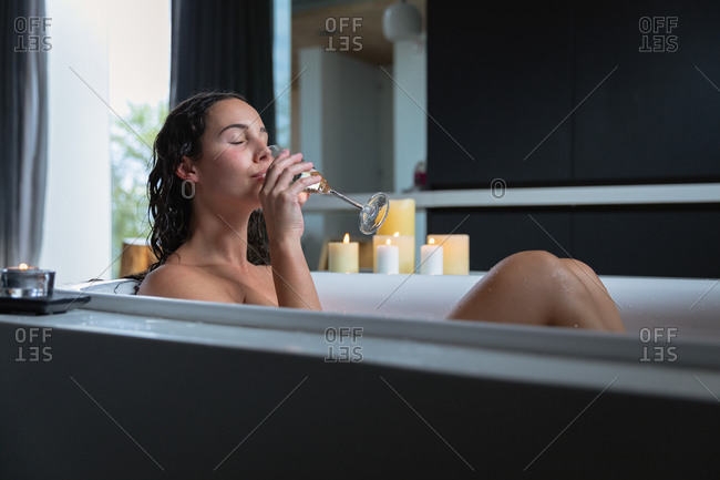 Side view of a young Caucasian brunette woman sitting in a bath with lit candles on the side, drinking champagne with her eyes closed