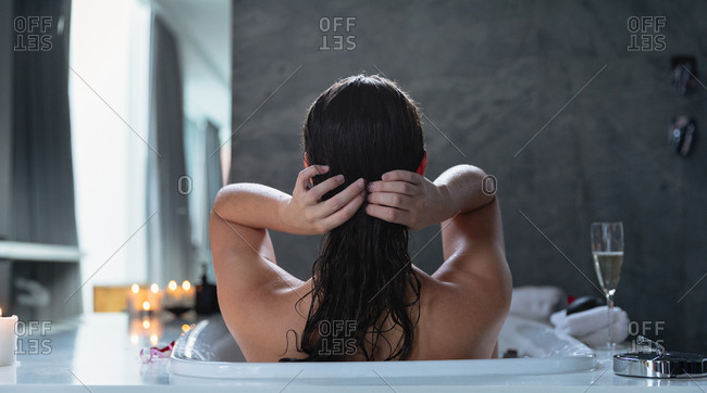 Rear view of a young Caucasian brunette woman sitting in a bath with lit candles and a glass of champagne on the side, adjusting her hair