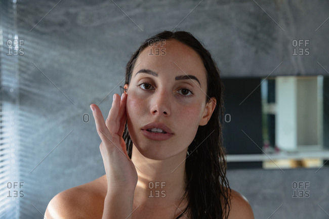 Portrait close up of a young Caucasian brunette woman looking straight to camera and massaging her face with one hand in a modern bathroom