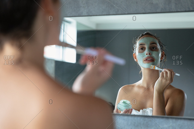 Over the shoulder view of a young Caucasian brunette woman wearing a bath towel looking in the mirror holding a jar and applying a face pack with a brush in a modern bathroom