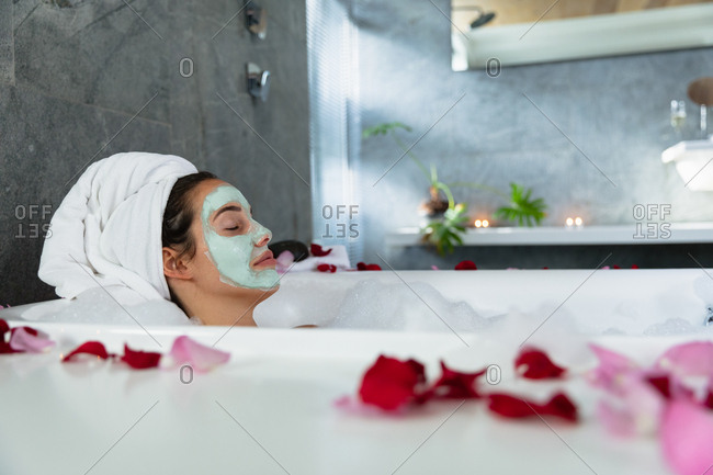 Side view of a young Caucasian brunette woman lying in a bath with lit candles and rose petals on the side, leaning back with her hair wrapped in a towel and her eyes closed