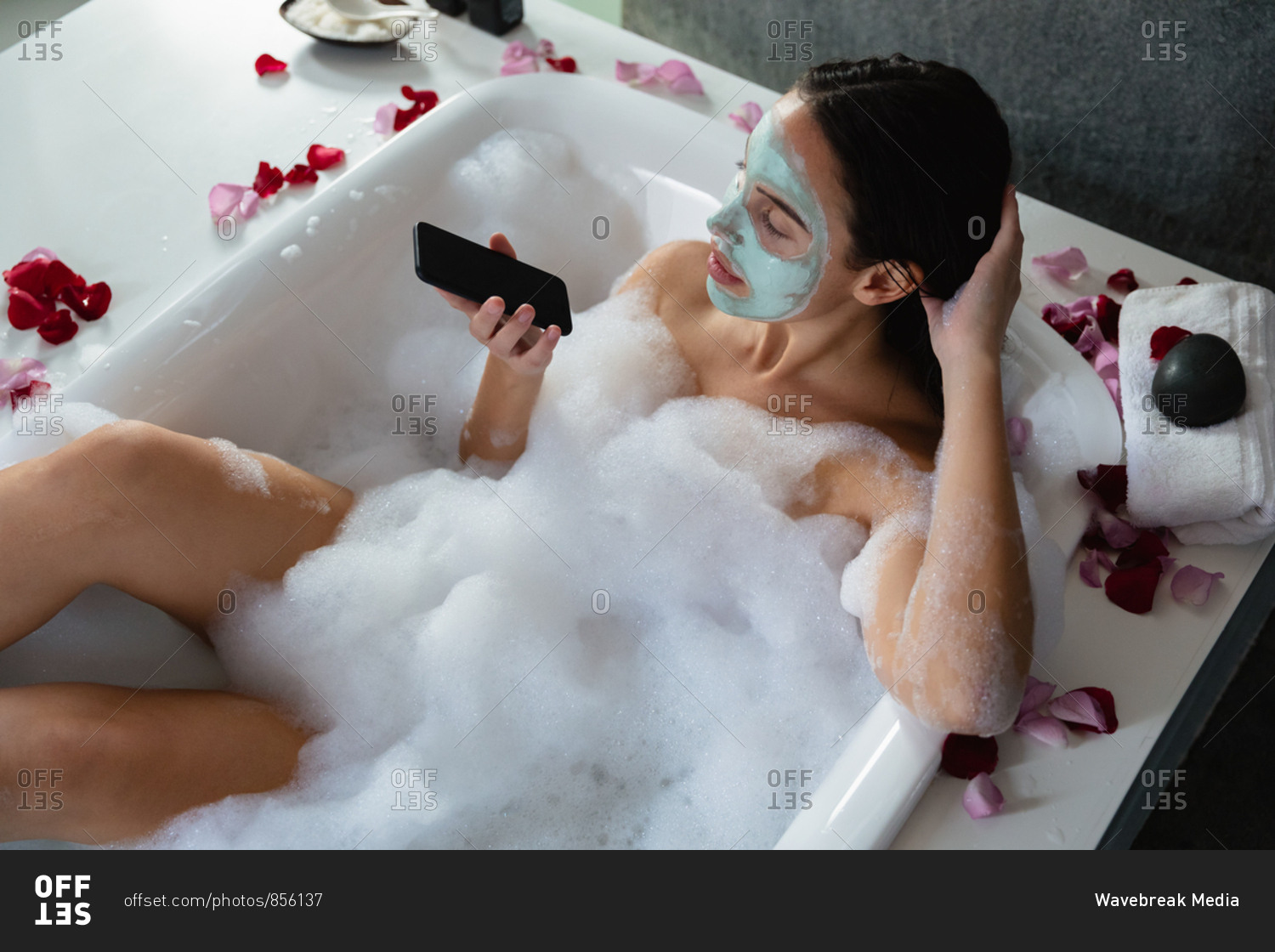 Elevated view of a young Caucasian woman wearing a face pack, sitting in a foam bath with rose petals around the edge using a smartphone