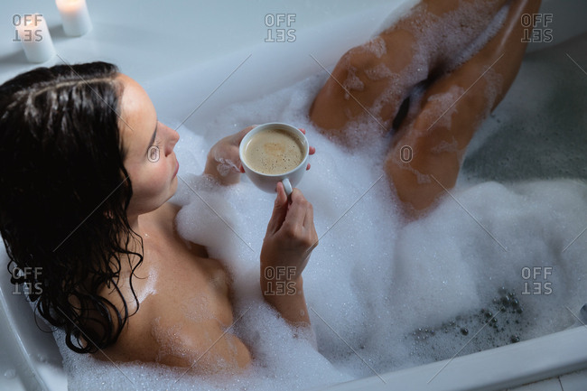 Elevated view of a young Caucasian brunette woman sitting in a foam bath with her feet up holding a cup of coffee