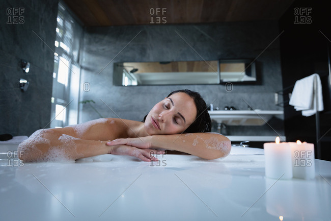 Portrait of a young Caucasian brunette woman sitting in a foam bath with lit candles on the edge, leaning on the side and resting with her eyes closed