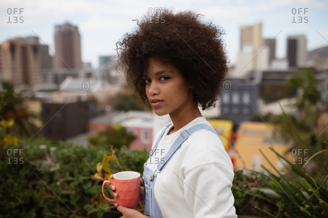 Portrait close up of a young mixed race woman standing outside on a balcony in the city holding a cup of coffee and turning her head to look to camera