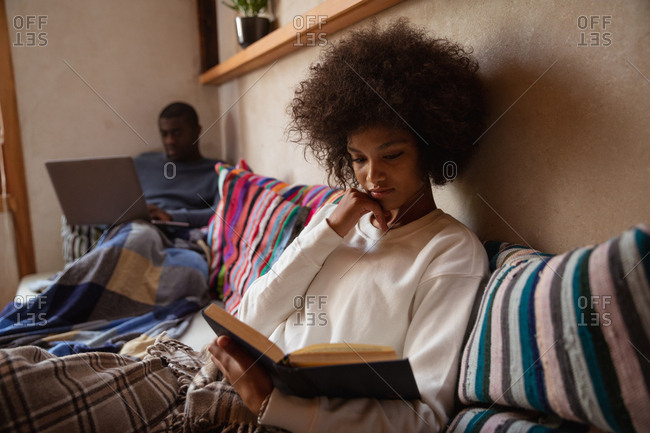 Front view close up of a young mixed race woman sitting on a sofa reading a book at home, her partner, a young African American man, is sitting on the sofa using a laptop computer in the background. They are both leaning on cushions and have blankets over their legs