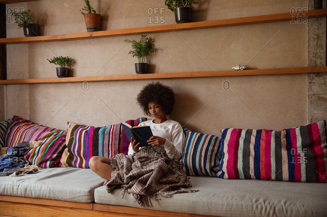 Front view of a young mixed race woman sitting on a sofa reading a book at home, leaning on cushions with a blanket over her legs
