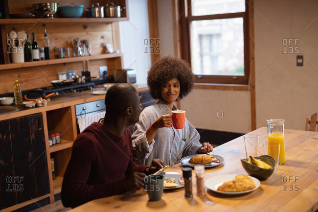 Front view of a smiling young mixed race woman and her partner, a young African American man, eating, talking and drinking coffee sitting at their kitchen table at home