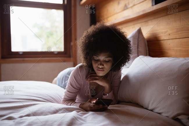 Front view close up of a young mixed race woman lying on her front on her bed leaning on her elbows using a smartphone at home