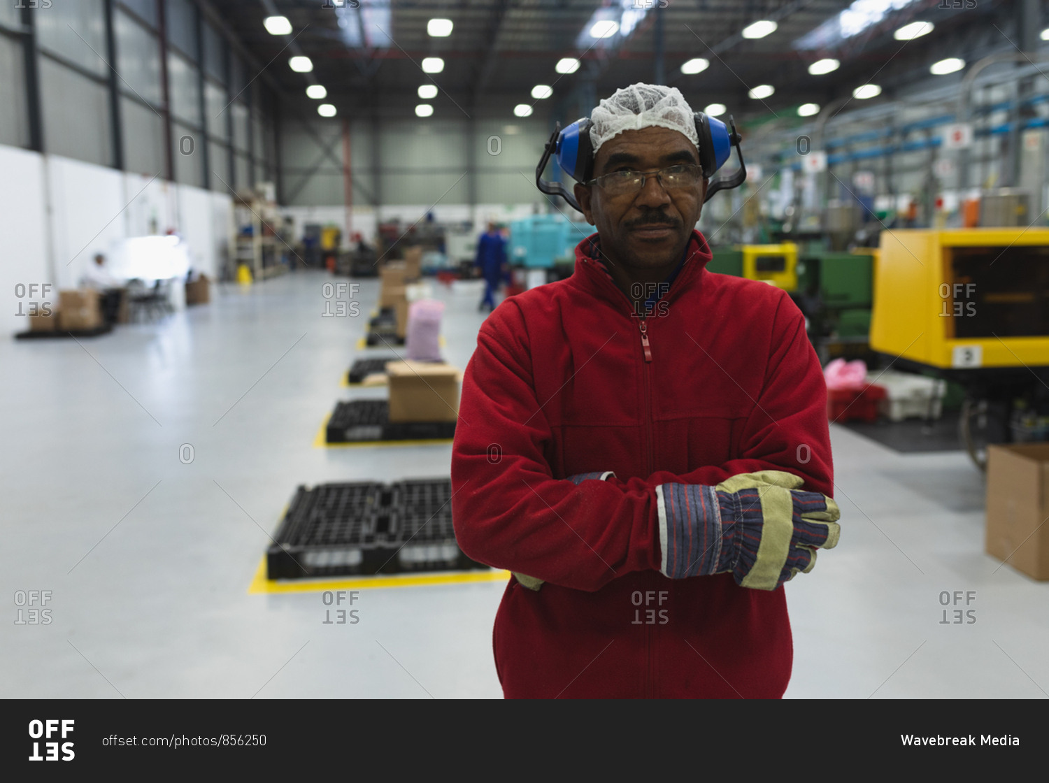 Portrait close up of a middle aged African American male factory worker dressed in workwear with ear defenders on his head, standing in a warehouse at a processing plant looking to camera with arms crossed