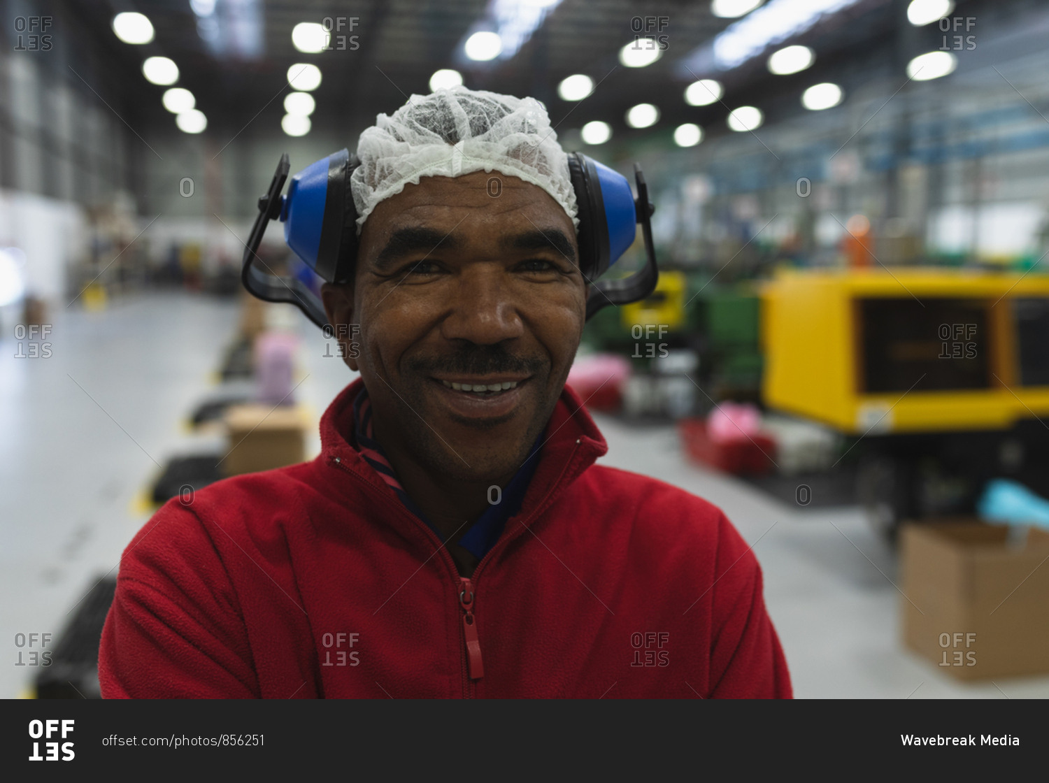 Portrait close up of a middle aged African American male factory worker dressed in workwear with ear defenders on his head, standing in a warehouse at a processing plant smiling to camera with arms crossed
