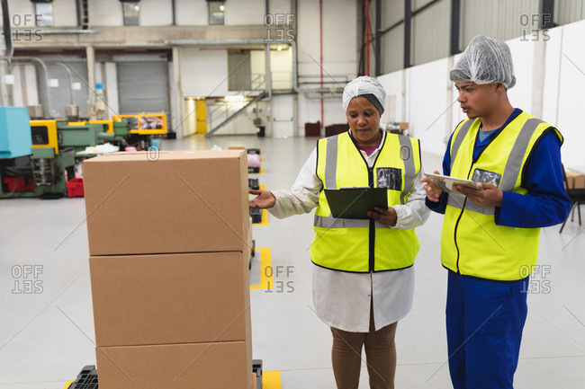 Front view close up of a young mixed race female and a young mixed race male factory worker talking in a warehouse loading bay at a factory. The woman is pointing to a stack of boxes and holding a clipboard, the man is using a tablet computer, both are dressed in workwear