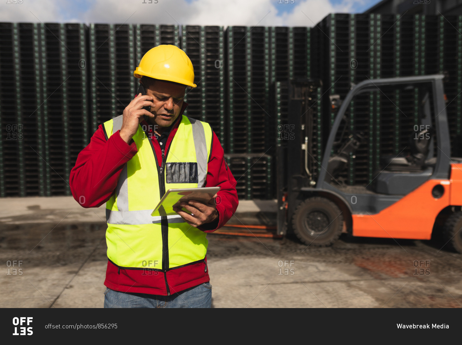 Front view close up of a middle aged Caucasian male factory worker on the phone using a tablet computer beside a forklift truck and stacks of pallets outside a warehouse at a factory