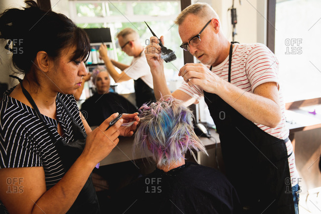 Side view of a middle aged Caucasian male hairdresser, a middle aged mixed race female hairdresser and a young Caucasian woman having her hair colored in a hair salon, reflected in a mirror