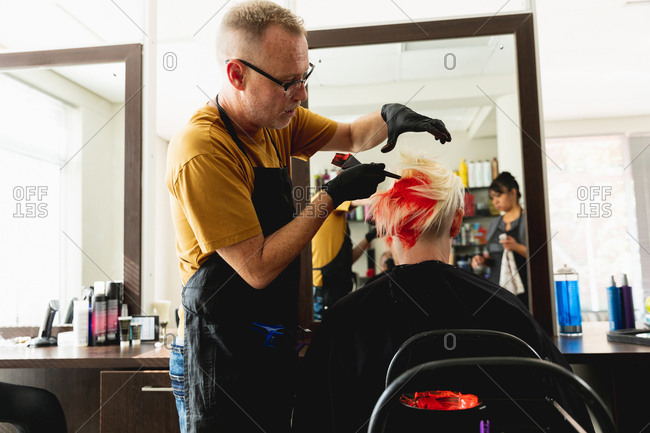 Middle aged Caucasian male hairdresser and a young Caucasian woman having her hair colored bright red in a hair salon and a middle aged mixed race female hairdresser reflected in a mirror