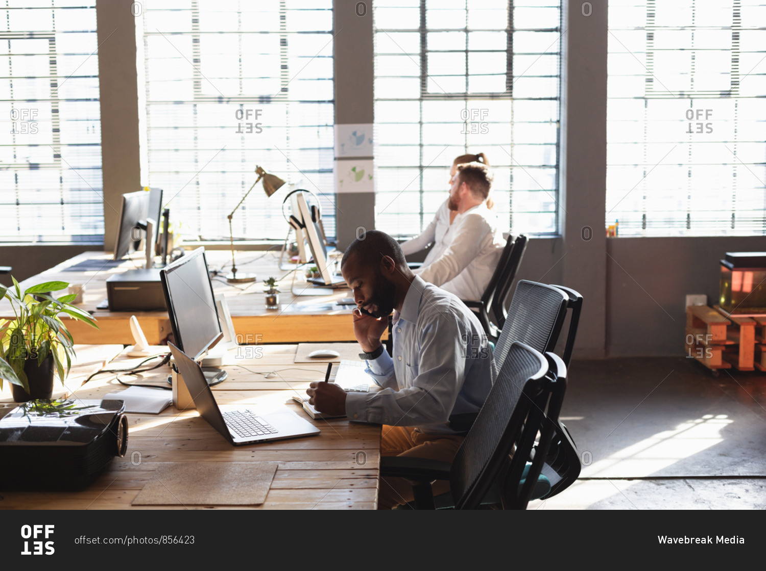 Side view of a young African American man sitting at a desk talking on ...