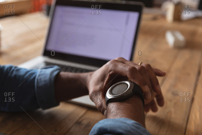 Close up of the hands of a young African American man sitting at a desk checking his watch and working on a laptop computer in a creative office
