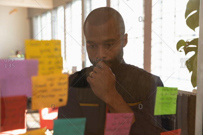 Front view close up of a young African American man reading notes on a glass wall and thinking during a team brainstorm session at a creative office, seen through glass wall