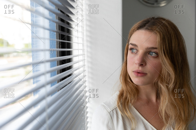 Front view close up of a young Caucasian woman standing with her head turned looking out of a window with venetian blinds in the modern office of a creative business