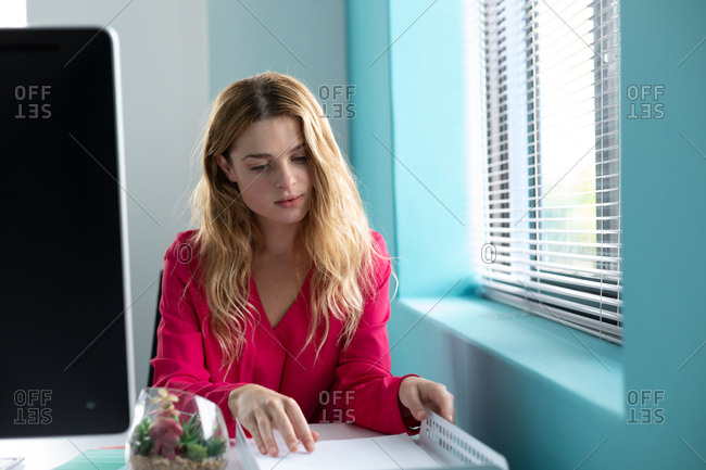 Front view close up of a young Caucasian woman sitting at a desk looking at paperwork in an in tray at the modern office of a creative business