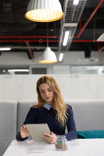 Front view of a young Caucasian woman sitting on a bench seat using a tablet computer at a table in the dining area of a creative business