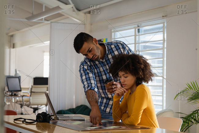 Side view of a young mixed race man standing and a young mixed race woman sitting at a desk having a discussion at a creative office