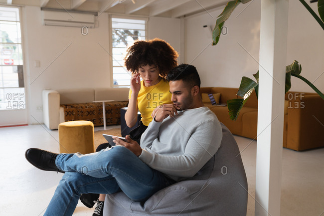 Front view of a young mixed race man and a young mixed race woman sitting on a bean bag using a tablet computer and having a discussion at a creative office