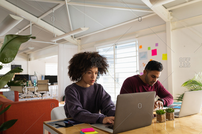 Front view of a young mixed race man and a young mixed race woman sitting at a desk using laptop computers at a creative office