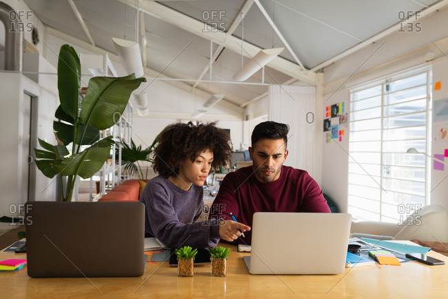Front view of a young mixed race man and a young mixed race woman sitting at a desk using laptop computers at a creative office