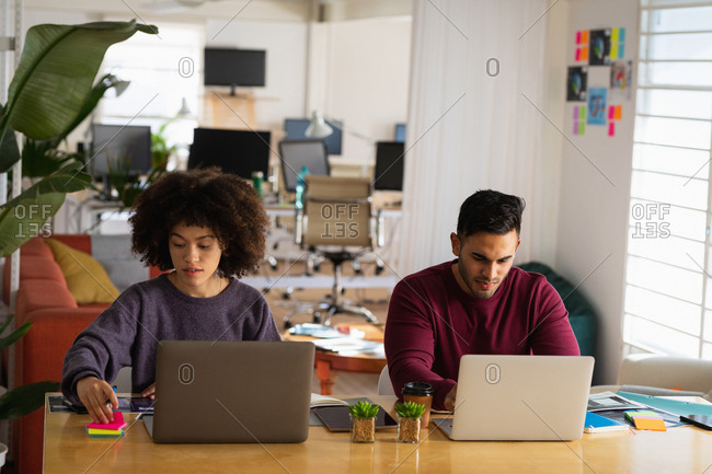 Front view of a young mixed race man and a young mixed race woman sitting at a desk using laptop computers at a creative office