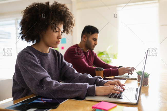 Side view close up of a young mixed race man and a young mixed race woman sitting at a desk using laptop computers at a creative office