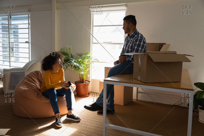 Side view close up of a young mixed race man sitting on a desk and a young mixed race woman sitting on a bean bag having a discussion with cardboard boxes at a creative office