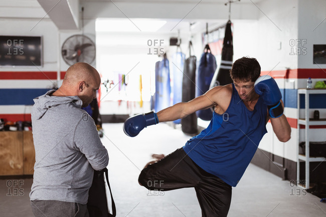 Front view of a young Caucasian male kickboxer practicing a kick against a pad held by a middle aged Caucasian male trainer in a boxing gym