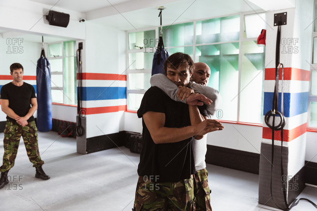 Front view of a middle aged Caucasian male instructor giving self defense training in a boxing gym demonstrating a hold on a young mixed race man, while another young man looks on