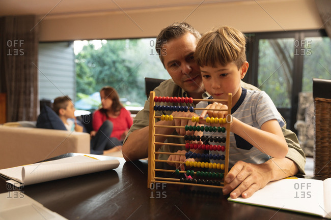 Front view of a middle aged Caucasian man helping his pre teen son to use abacus, the mother talking with their other son in the background